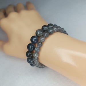 May include: Two beaded bracelets with dark grey and blue-grey spherical beads. The bracelets are worn on a wrist. The beads have a polished appearance and vary in size, creating a layered effect. The background is a soft white.