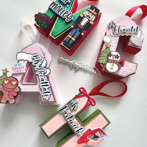 May include: A collection of personalized Christmas ornaments shaped like letters. Each ornament features a name, decorative elements like a snowman, gingerbread man, or nutcracker, and a ribbon for hanging. The ornaments are in various colors, including red, pink, and green.