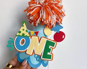 May include: A light blue party hat featuring a large orange and white yarn pom-pom on top. The front displays the word ONE in blue, yellow, and green letters with star patterns, accented by a small green party hat, colorful bunting, and balloons.