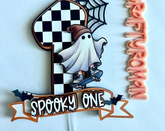 May include: A Halloween-themed cake topper featuring a number one with a black and white checkered pattern. A ghost wearing a beanie rides a skateboard. The words "Spooky One" are on a banner with bats. The text "CRAFTY WOMAN" is also present.
