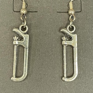 May include: A pair of silver-toned dangle earrings featuring a miniature hacksaw charm. The earrings are attached to a gray display card and include standard silver-colored metal fishhook ear wires with a small bead accent above the saw charm.