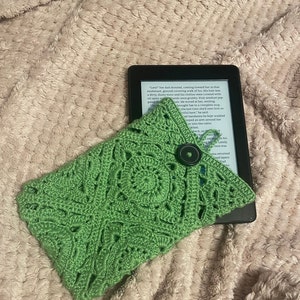 May include: A green crocheted e-reader cover with a black button. The cover is made with a square stitch pattern.