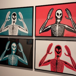 May include: Four framed art prints featuring a stylized skeleton figure with arms raised. Each print has a different background color: teal, red, black, and white. The skeleton is white or red with black details.