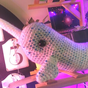 May include: A crocheted narwhal toy, predominantly white with blue spots and a long tusk. The toy has a black eye and is positioned on a small wooden structure. The craft item is likely handmade and intended for decorative purposes.