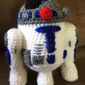 May include: A crocheted R2-D2 robot toy with white, gray, blue, and red details. The toy has a black eye and a red button on its head.