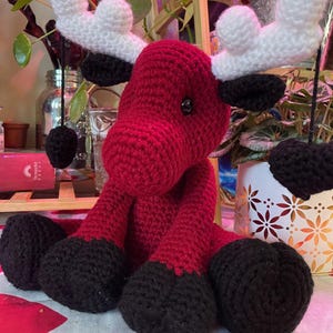 Może przedstawiać: Ręcznie robiony szydełkowy łoś z czerwonym korpusem, czarnymi kopytami i białymi rogami. Łoś amigurumi ma okrągłą głowę i siedzi. Łoś to miękka, pluszowa zabawka.