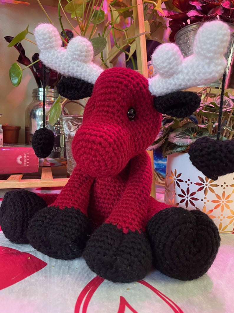 Może przedstawiać: Ręcznie robiony szydełkowy łoś z czerwonym korpusem, czarnymi kopytami i białymi rogami. Łoś amigurumi ma okrągłą głowę i siedzi. Łoś to miękka, pluszowa zabawka.