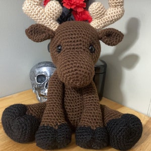 May include: A brown crocheted moose with black hooves, sitting on a wooden surface. The moose has beige antlers and black eyes.