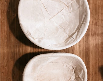 May include: Two off-white, round and oval-shaped bread proofing baskets. The baskets are made of a light-colored fabric and are set against a wooden background. The round basket is at the top, and the oval basket is below.