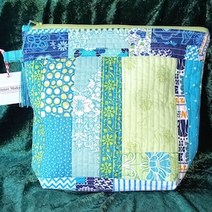 May include: A patchwork zipped pouch in shades of blue, teal, and green. The bag features various fabric patterns, including polka dots, floral designs, and geometric shapes. A tag with the text "Sister Make" is attached to the zipper pull.