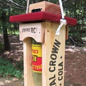 May include: Wooden bird feeder with a red roof and a glass bottle insert. The feeder is hanging from a rope and has the text "Drink RC/ Royal Crown Cola" printed on it.