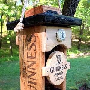 Pode incluir: Um comedouro para p&aacute;ssaros de madeira em forma de garrafa de cerveja Guinness. O comedouro &eacute; pintado de preto e marrom e tem uma pequena bandeja para sementes de p&aacute;ssaros. O comedouro est&aacute; pendurado em um galho de &aacute;rvore.