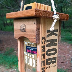 May include: A hanging bird feeder constructed from wood and a glass Knob Creek bourbon bottle. The wooden frame features the text Knob Creek, barrel illustrations, and a circular stamp. A small wooden tray is attached at the bottom for bird seed.