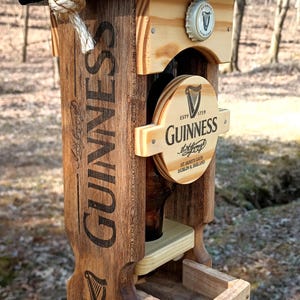 K&ouml;nnte beinhalten: Ein h&ouml;lzerner Vogelh&auml;uschen in Form einer Guinness-Bierflasche. Der Futterspender hat ein schwarzes Oberteil und eine braune Holzbasis mit dem aufgedruckten Wort "GUINNESS". Der Futterspender hat eine kleine Schale f&uuml;r Vogelfutter und eine klare Glasflasche f&uuml;r Wasser.