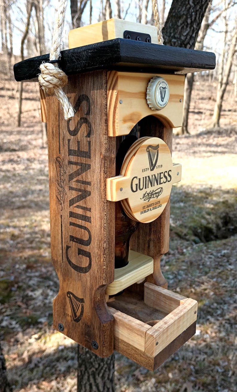 K&ouml;nnte beinhalten: Ein h&ouml;lzerner Vogelh&auml;uschen in Form einer Guinness-Bierflasche. Der Futterspender hat ein schwarzes Oberteil und eine braune Holzbasis mit dem aufgedruckten Wort "GUINNESS". Der Futterspender hat eine kleine Schale f&uuml;r Vogelfutter und eine klare Glasflasche f&uuml;r Wasser.