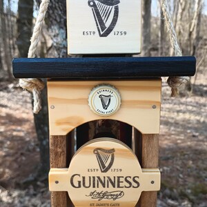 K&ouml;nnte beinhalten: Holzwindspiel mit einer Guinness-Bierflasche mit einem schwarz-wei&szlig;en Logo und dem Text "ESTD 1759 GUINNESS Arthur Guinness St. James's Gate Dublin 8, Ireland". Das Windspiel h&auml;ngt an einem Seil und hat eine schwarze Oberseite.