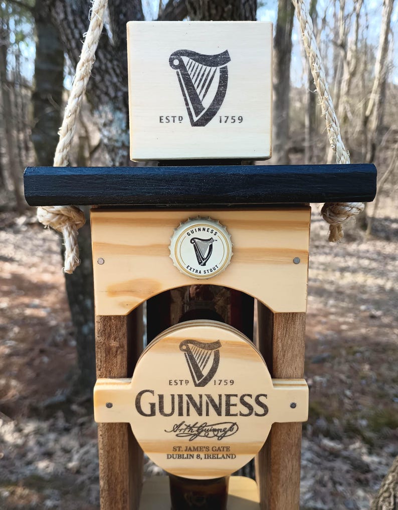 K&ouml;nnte beinhalten: Holzwindspiel mit einer Guinness-Bierflasche mit einem schwarz-wei&szlig;en Logo und dem Text "ESTD 1759 GUINNESS Arthur Guinness St. James's Gate Dublin 8, Ireland". Das Windspiel h&auml;ngt an einem Seil und hat eine schwarze Oberseite.