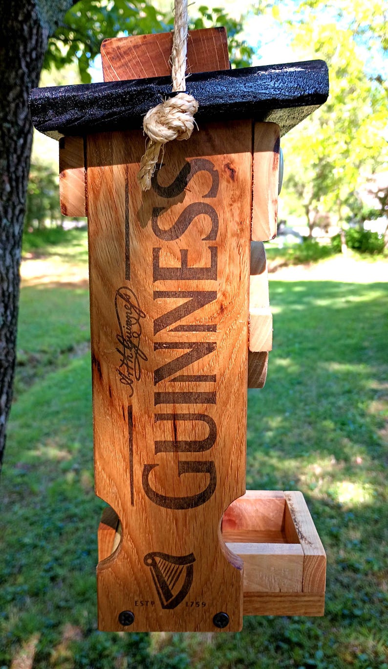 Pode incluir: Um alimentador de p&aacute;ssaros de madeira com um topo preto e uma corda para pendurar. O alimentador &eacute; decorado com o logotipo da Guinness e o texto "Arthur Guinness, Dublin" e "Est. 1759".