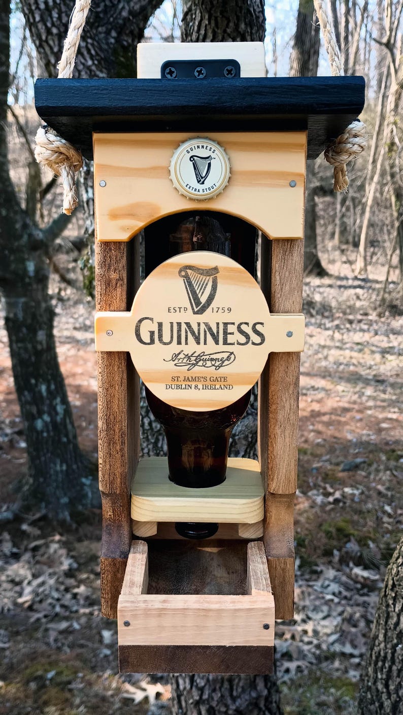 K&ouml;nnte beinhalten: Vogelhaus aus Holz mit einer wiederverwendeten Guinness-Bierflasche. Der Futterspender hat ein schwarzes Dach und eine braune Basis. Das Guinness-Logo ist auf der Flasche und dem Futterspender sichtbar.
