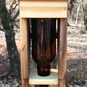 K&ouml;nnte beinhalten: Ein h&ouml;lzerner Vogelh&auml;uschen mit schwarzem Oberteil und einer braunen Glasflasche im Inneren. Der Futterspender h&auml;ngt an einem Seil und tr&auml;gt ein Schild mit der Aufschrift "Sunny Side Workshop".