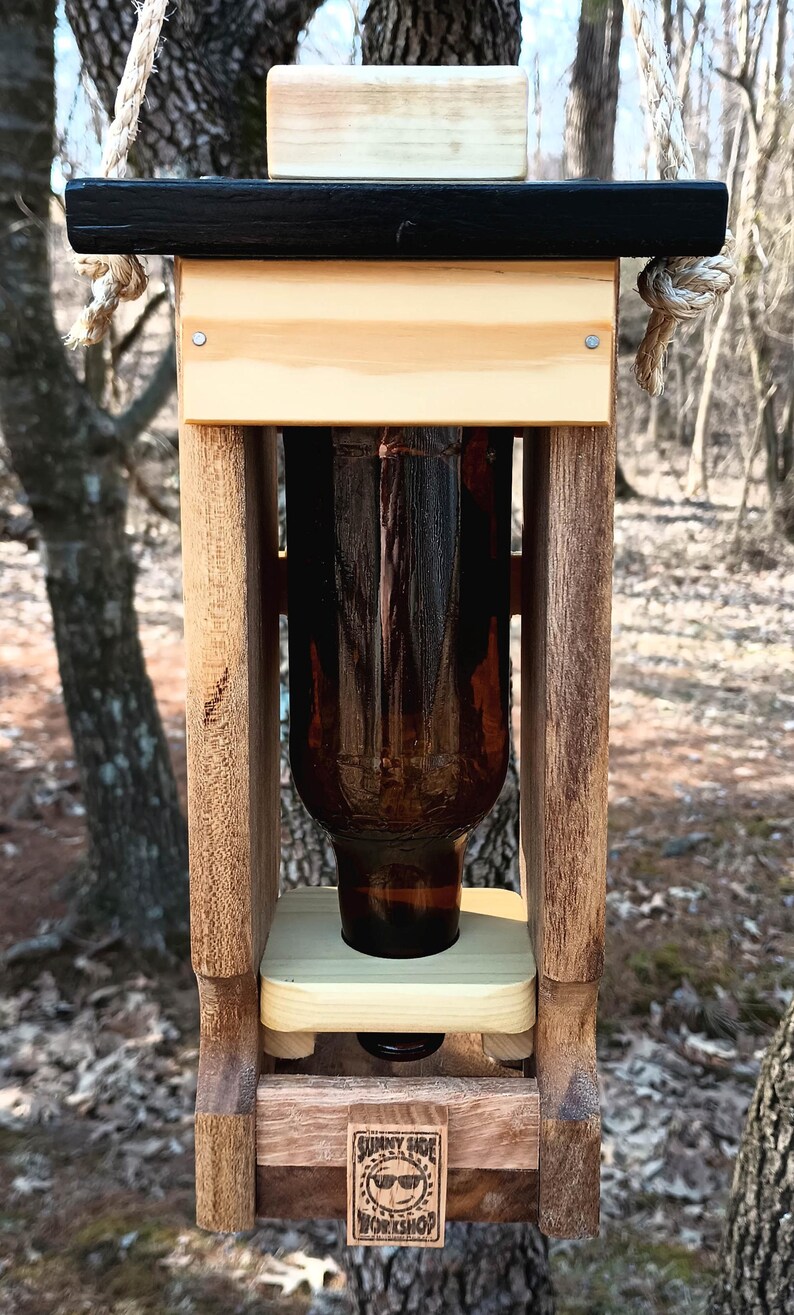 K&ouml;nnte beinhalten: Ein h&ouml;lzerner Vogelh&auml;uschen mit schwarzem Oberteil und einer braunen Glasflasche im Inneren. Der Futterspender h&auml;ngt an einem Seil und tr&auml;gt ein Schild mit der Aufschrift "Sunny Side Workshop".