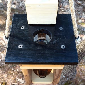 K&ouml;nnte beinhalten: Ein Vogelh&auml;uschen aus Holz mit schwarzem Oberteil und einer durchsichtigen Glasflasche im Inneren. Der Futterspender ist an einem Seil aufgeh&auml;ngt und hat einen Holzdeckel oben.