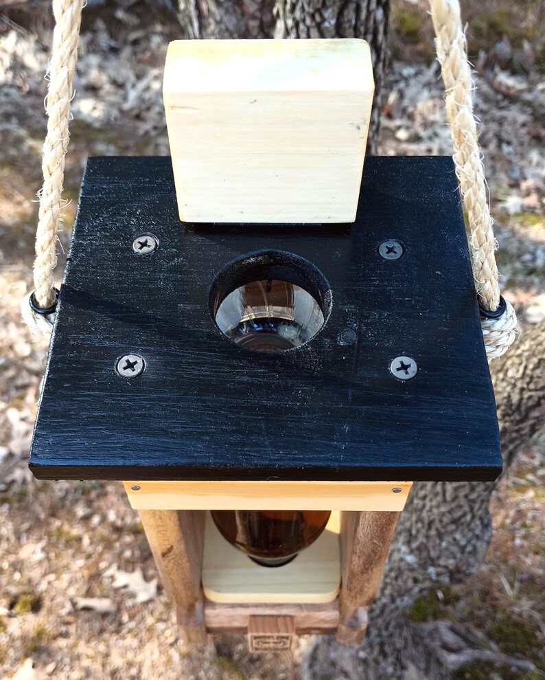 K&ouml;nnte beinhalten: Ein Vogelh&auml;uschen aus Holz mit schwarzem Oberteil und einer durchsichtigen Glasflasche im Inneren. Der Futterspender ist an einem Seil aufgeh&auml;ngt und hat einen Holzdeckel oben.