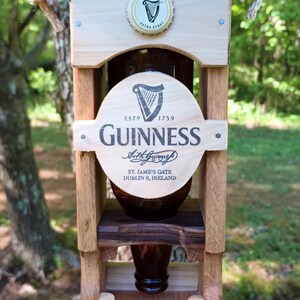 Pode incluir: Um alimentador de p&aacute;ssaros de madeira em forma de casa com uma garrafa de Guinness no centro. A garrafa &eacute; cercada por uma moldura de madeira com o logotipo e o texto da Guinness. O alimentador tem um telhado preto e uma base de madeira.