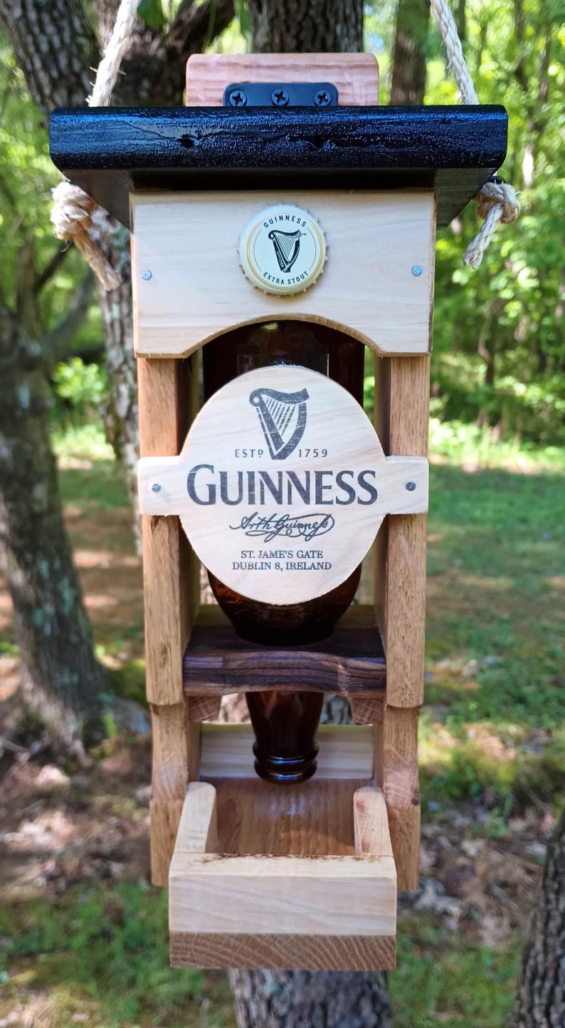 Pode incluir: Um alimentador de p&aacute;ssaros de madeira em forma de casa com uma garrafa de Guinness no centro. A garrafa &eacute; cercada por uma moldura de madeira com o logotipo e o texto da Guinness. O alimentador tem um telhado preto e uma base de madeira.