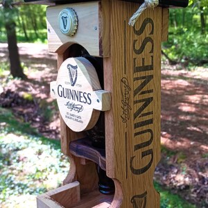Pode incluir: Um comedouro para p&aacute;ssaros de madeira em forma de garrafa de cerveja Guinness. O comedouro tem um telhado preto e uma pequena bandeja de madeira para sementes de p&aacute;ssaros. A garrafa &eacute; etiquetada com o logotipo da Guinness e o texto "Est. 1759".