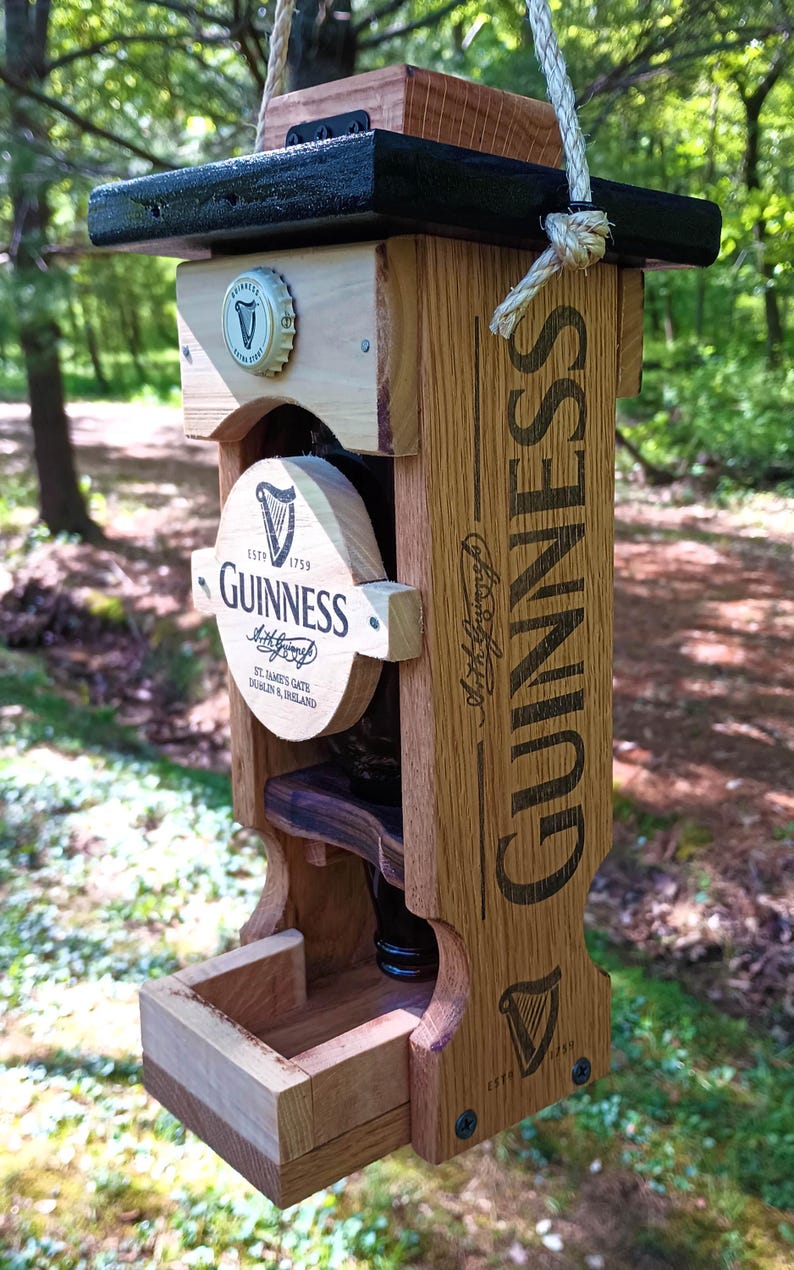 Pode incluir: Um comedouro para p&aacute;ssaros de madeira em forma de garrafa de cerveja Guinness. O comedouro tem um telhado preto e uma pequena bandeja de madeira para sementes de p&aacute;ssaros. A garrafa &eacute; etiquetada com o logotipo da Guinness e o texto "Est. 1759".