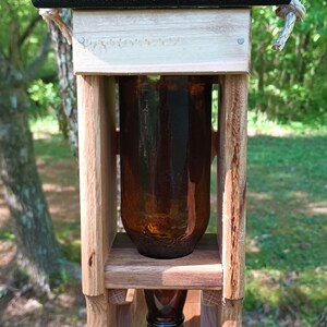 Pode incluir: Um alimentador de p&aacute;ssaros de madeira com uma garrafa de vidro marrom pendurada em uma corda. O alimentador tem um topo pintado de preto e uma placa de madeira que diz "Sunny Side Workshop".