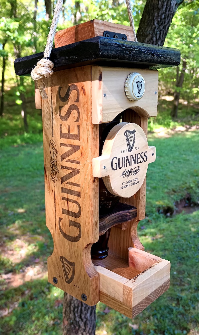 Pode incluir: Um comedouro para p&aacute;ssaros de madeira em forma de garrafa de cerveja Guinness. O comedouro &eacute; pintado de preto e marrom e tem uma pequena bandeja para sementes de p&aacute;ssaros. O comedouro est&aacute; pendurado em um galho de &aacute;rvore.