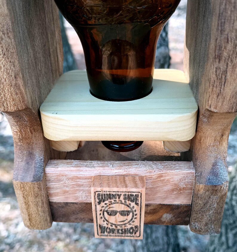 K&ouml;nnte beinhalten: Ein h&ouml;lzerner Vogelh&auml;uschen mit einer Glasflasche, die oben befestigt ist. Der Futterspender hat eine kleine Plattform, auf der die Flasche stehen kann, und ein Schild mit der Aufschrift "Sunny Side Workshop".
