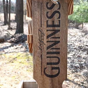 K&ouml;nnte beinhalten: Vogelhaus aus Holz mit schwarzem Oberteil und Guinness-Logo. Der Futterspender hat eine kleine Schale f&uuml;r Vogelfutter und ein Seil zum Aufh&auml;ngen.