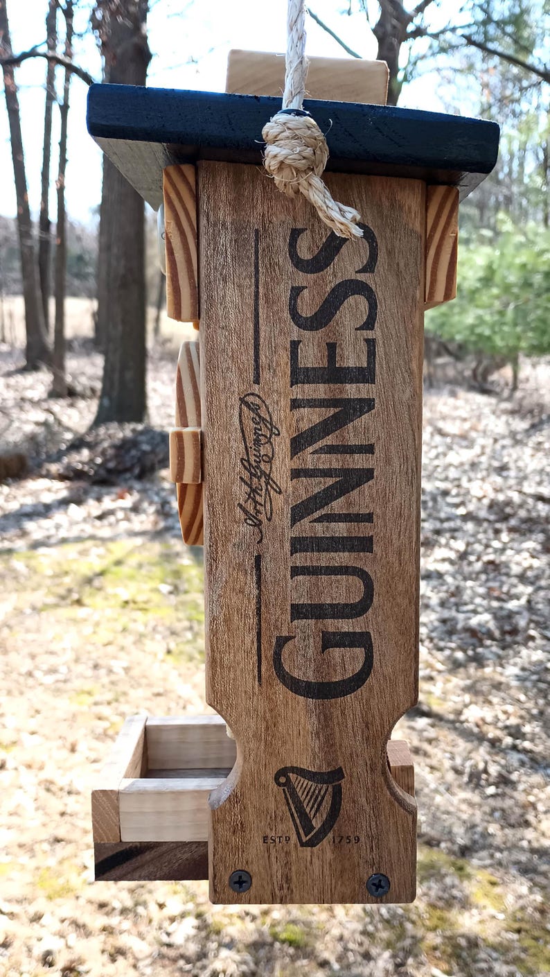 K&ouml;nnte beinhalten: Vogelhaus aus Holz mit schwarzem Oberteil und Guinness-Logo. Der Futterspender hat eine kleine Schale f&uuml;r Vogelfutter und ein Seil zum Aufh&auml;ngen.