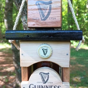 Pode incluir: Abridor de garrafas de madeira com uma parte superior de metal preto e uma tampa de garrafa de Guinness. O abridor de garrafas tem um logotipo da Guinness com o texto "ESTD 1759 GUINNESS" e as palavras "Arthur Guinness" em it&aacute;lico.