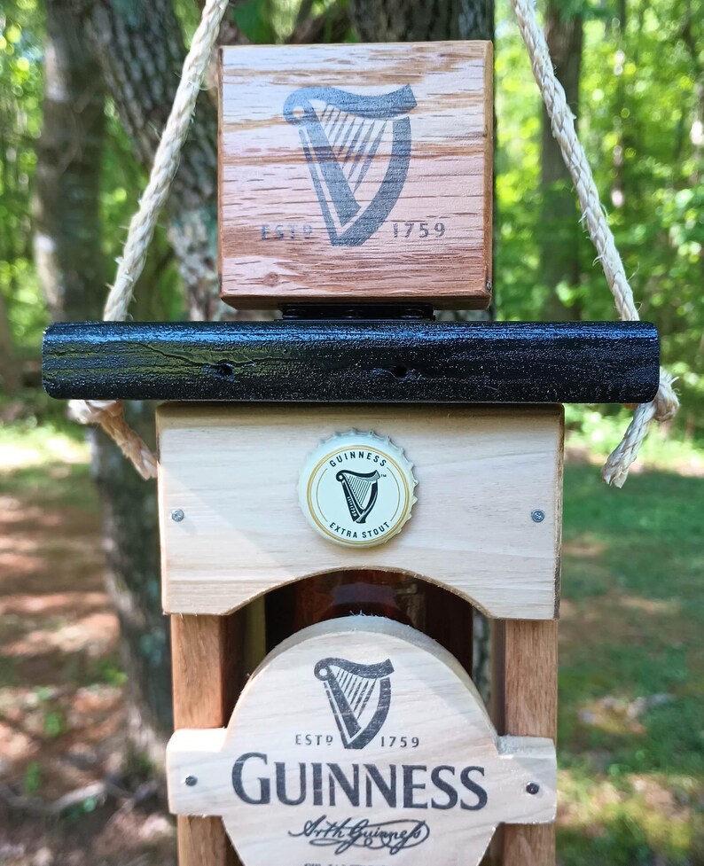 Pode incluir: Abridor de garrafas de madeira com uma parte superior de metal preto e uma tampa de garrafa de Guinness. O abridor de garrafas tem um logotipo da Guinness com o texto "ESTD 1759 GUINNESS" e as palavras "Arthur Guinness" em it&aacute;lico.