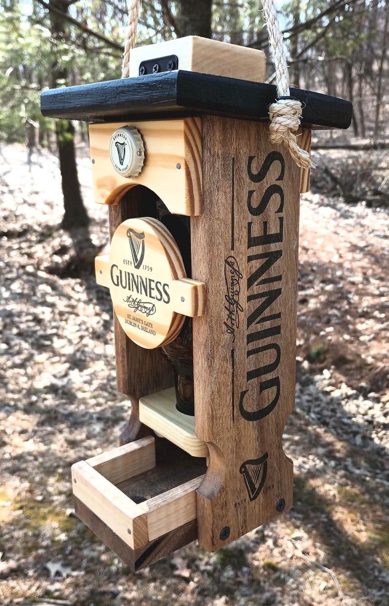 K&ouml;nnte beinhalten: Ein h&ouml;lzerner Vogelh&auml;uschen in Form einer Guinness-Bierflasche. Der Futterspender hat ein schwarzes Oberteil, eine klare Glasflasche und eine Holzschale f&uuml;r Vogelfutter. Der Futterspender h&auml;ngt an einem Seil.