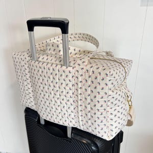 Può includere: Una borsa da viaggio trapuntata bianca con un motivo floreale di fiori blu e rossi. La borsa ha una chiusura a cerniera, una tracolla ed è posizionata sopra una valigia nera con ruote con il manico esteso.