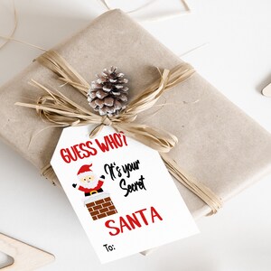 May include: White gift tag with red and black text that says "Guess Who? It's Your Secret Santa" with a cartoon image of Santa Claus climbing down a chimney. There is a line at the bottom that says "To:"