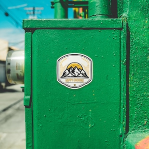 May include: A green metal electrical box with a white sticker featuring a yellow sun, black mountains, and the text "Happy Caching".