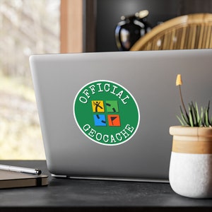 May include: A green circle sticker with white text that says "OFFICIAL GEOCACHE" and a compass rose with a person icon in the center. The sticker is on a silver laptop.