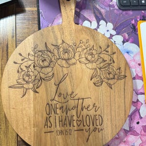 May include: Wooden serving board with a handle, featuring engraved floral designs and the text "Love one another as I have loved you" from John 15:12. The board is round with a light brown finish.