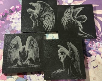 Puede incluir: Conjunto de cinco posavasos cuadrados de pizarra negra. Cada posavasos presenta una imagen grabada de un ángel con grandes alas. Las figuras de ángeles están en varias poses, creando una estética gótica o vintage.