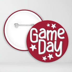 May include: Two round buttons. One is white with a red border and a pin back. The other is red with white text that reads "Game Day" and white star accents. The buttons are likely for a sports or event.