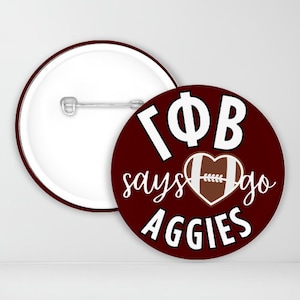 May include: A maroon button with white text that reads "Gamma Phi Beta says go Aggies" with a white outline and a football-shaped heart in the center.