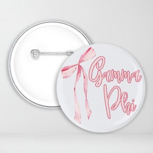 May include: A white button with a pink bow and the text "Gamma Phi" in pink lettering.