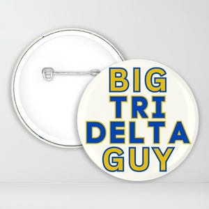 May include: A white button with a blue and yellow design. The text on the button reads "BIG TRI DELTA GUY".