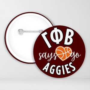 May include: A maroon button with white text that reads "Gamma Phi Beta says go Aggies" with an orange basketball in a heart shape.
